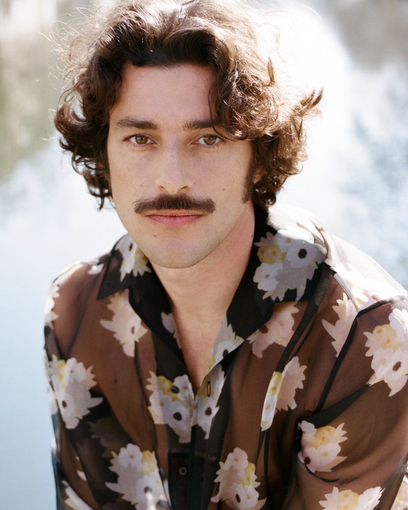 Portrait of a man with a mustache wearing a floral-patterned shirt, posing outdoors with soft, diffused natural lighting.