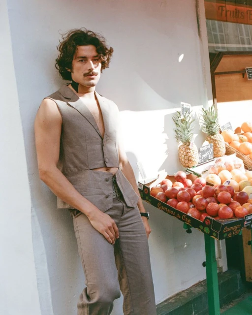Man in vest and pants poses next to fruit stand. He has dark curly hair and a mustache. Image shows a fashion or lifestyle shot.