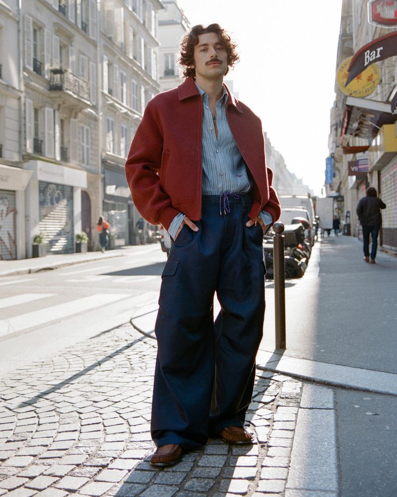 A stylish adult male with a mustache poses on a cobblestone street in a red jacket, blue shirt, and baggy pants with city buildings.