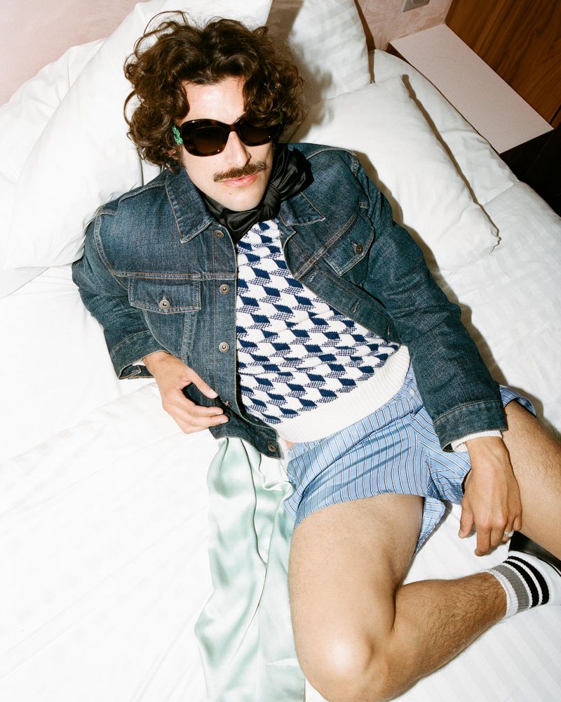 Man wearing sunglasses and a denim jacket reclining on a white bed for Man About Town.