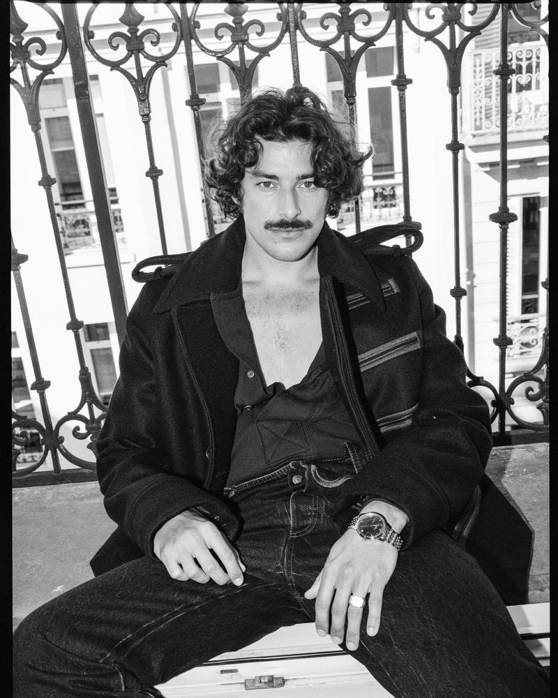 Black and white portrait of a man with a mustache sitting on a bench, wearing a jacket and jeans in front of a building.