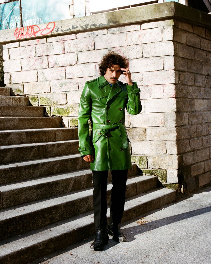 An adult man stands on concrete stairs outside, wearing a green leather trench coat, black pants, and dark shoes. The background is a brick wall.