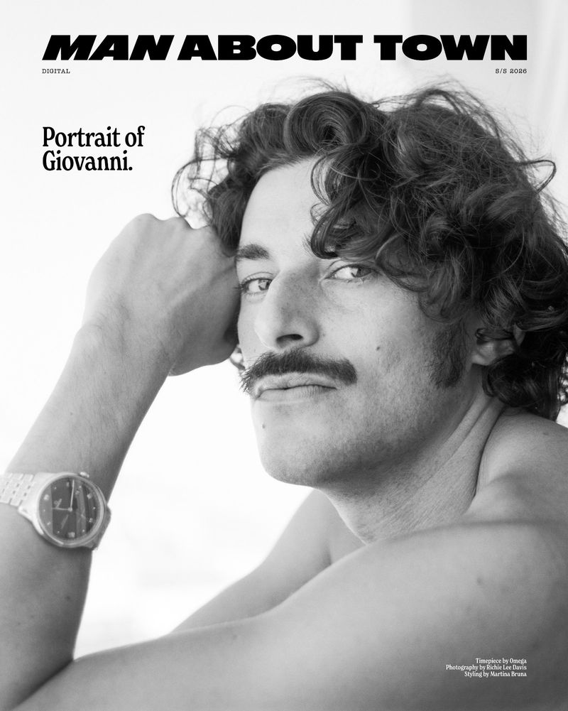Portrait of a man with dark curly hair and a mustache wearing a silver wristwatch against a bright white background.