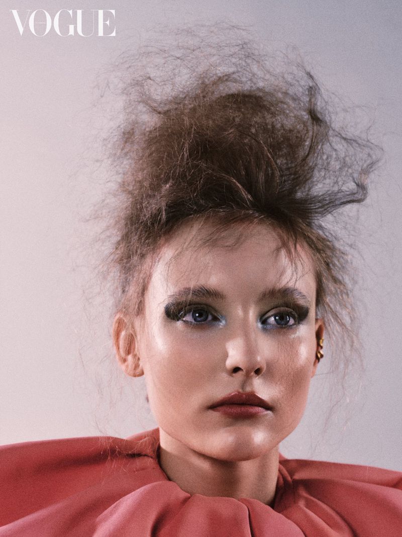 Close-up beauty portrait of a woman with dramatic eye makeup, textured updo hair, and a pink ruffled collar against a pale background