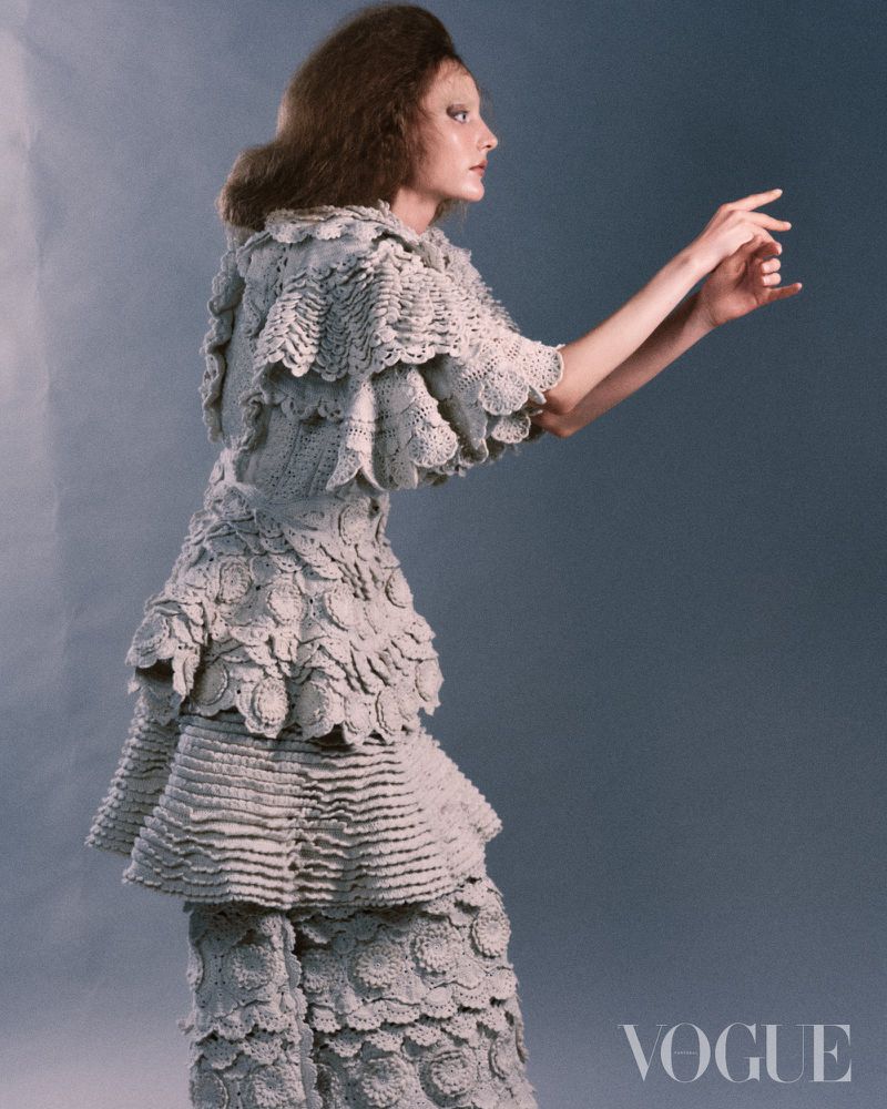 Model in textured grey ruffled crochet outfit gesturing forward against a grey studio backdrop