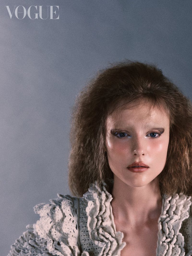 Close-up portrait of a woman with voluminous textured hair and decorative knitwear against a grey backdrop for Vogue Portugal.