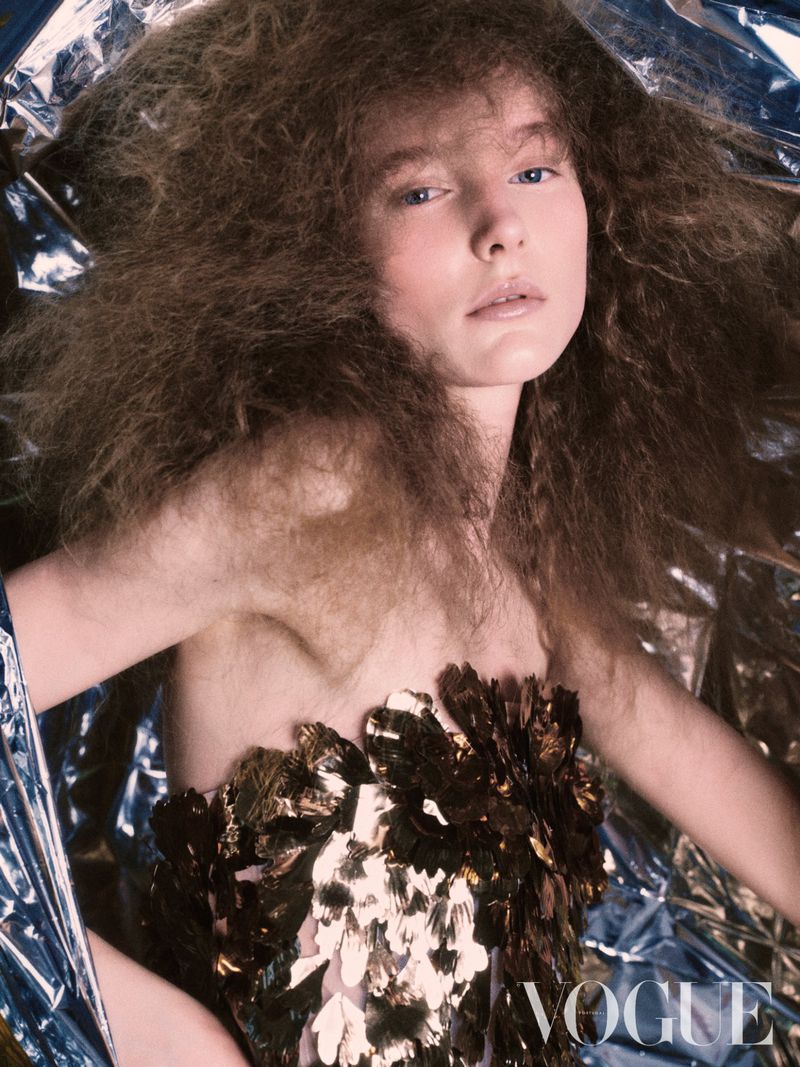 Fashion shot of a young woman with curly brown hair wearing a gold-colored bodice against a shiny, crinkled silver foil background.