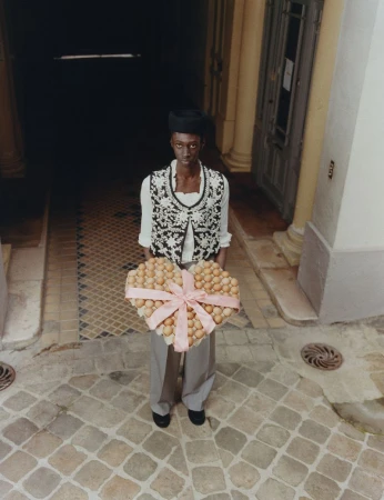 Model standing on cobblestone holding a large heart-shaped arrangement of eggs tied with a pink ribbon for Luncheon Magazine SS26.