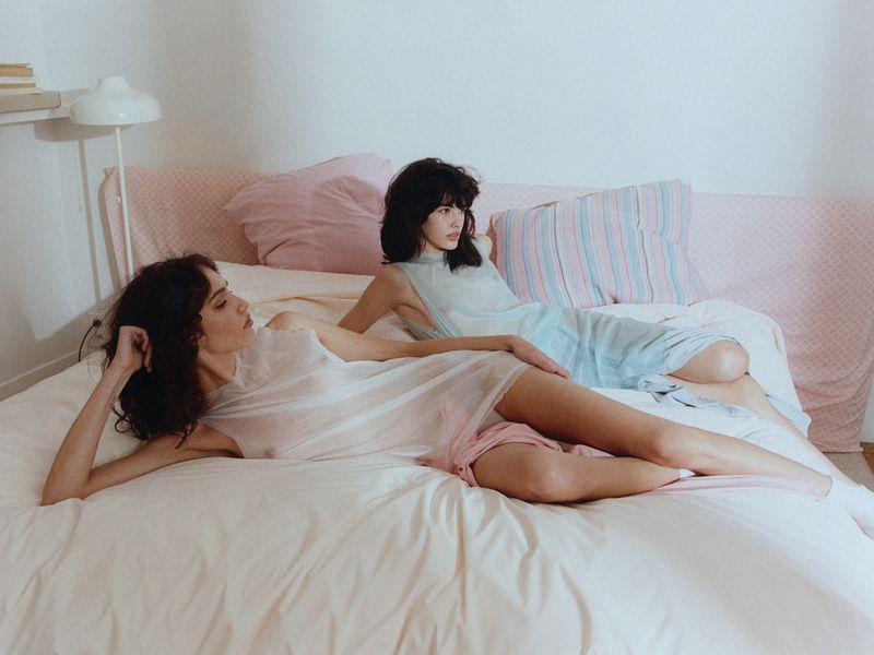 Two women lying on a large white bed wearing sheer sleeveless dresses in a domestic bedroom setting.