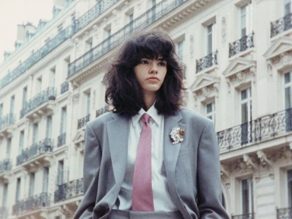 Model in a grey oversized suit jacket, white shirt, and pink tie standing before a Haussmann building exterior