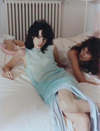 Two women lying on a rumpled white bed in a bedroom featuring a radiator and soft natural light.
