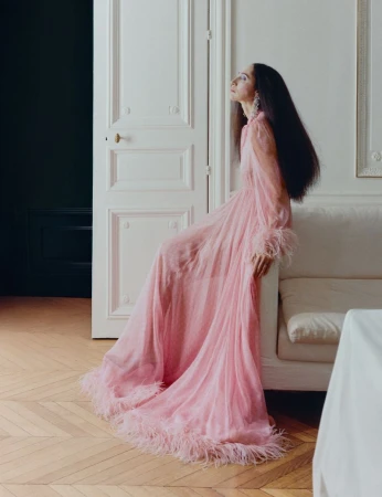 Model with long dark hair wearing a long pink sheer dress sitting on a white sofa in a room with paneled doors.