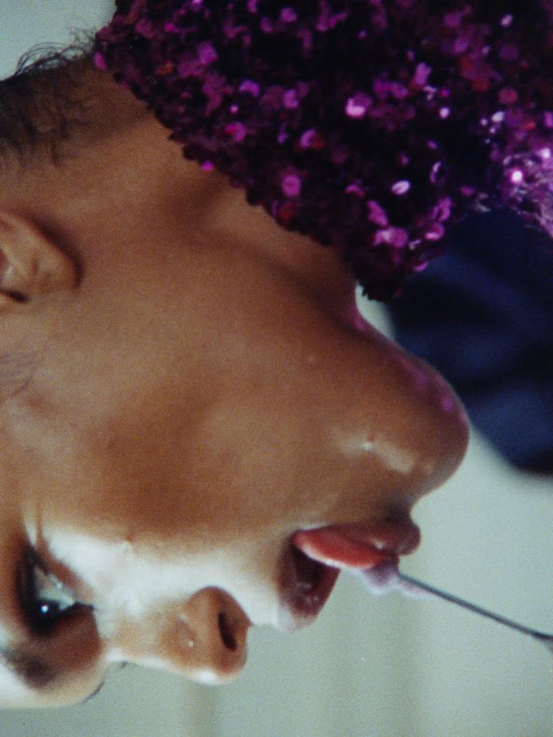Close-up profile of a model wearing a purple sequin garment while licking a spoon with cream.
