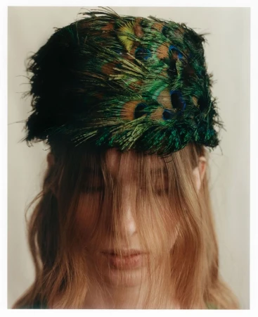Model wearing a structured peacock feather pillbox hat with hair falling across face for Vogue Polska Different Age editorial
