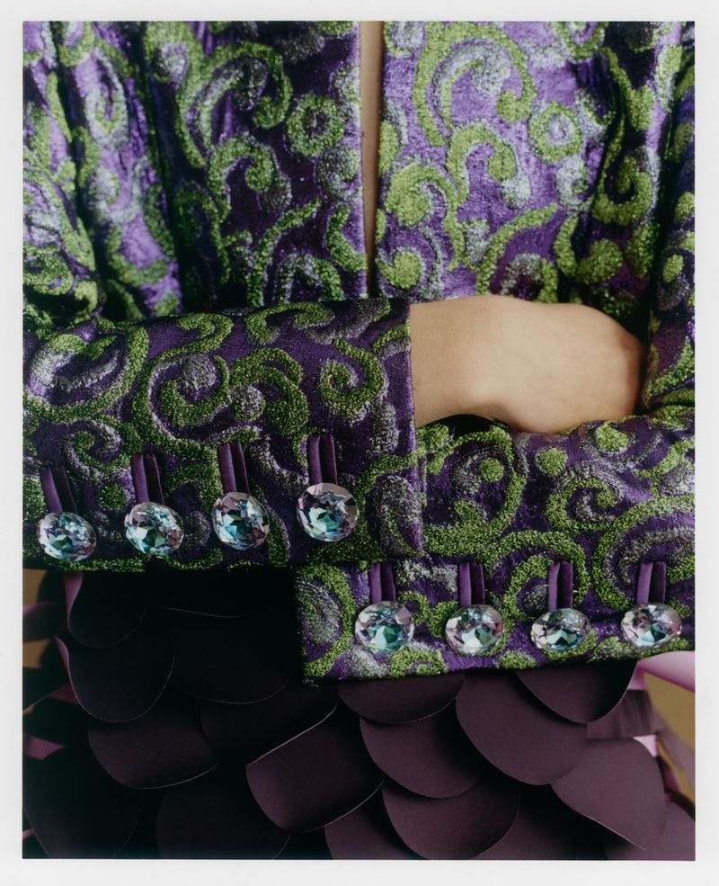 Close-up of a person wearing a purple and green embroidered patterned jacket with jeweled buttons and layered dark skirt.