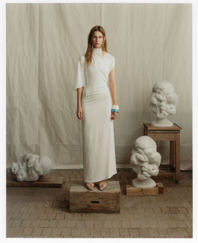 Model standing on a wooden crate wearing a long white dress in an artist studio with white fabric backdrop and plaster sculptures