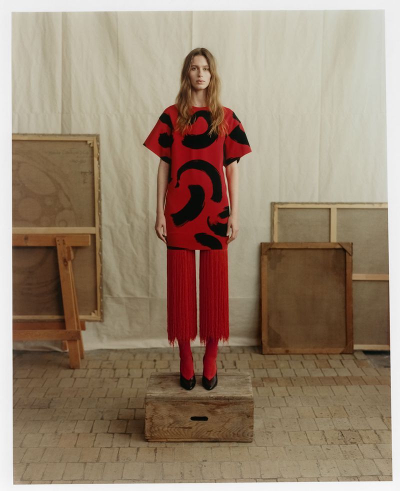 Model standing on a wooden crate wearing a red patterned top and red fringed trousers in an artist studio.