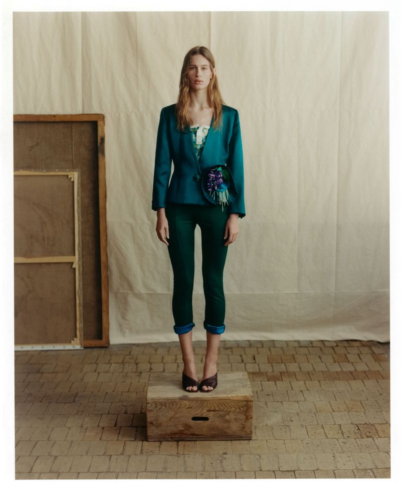 Model standing on a wooden crate wearing a teal blazer and dark green trousers against a cream canvas backdrop in a studio.