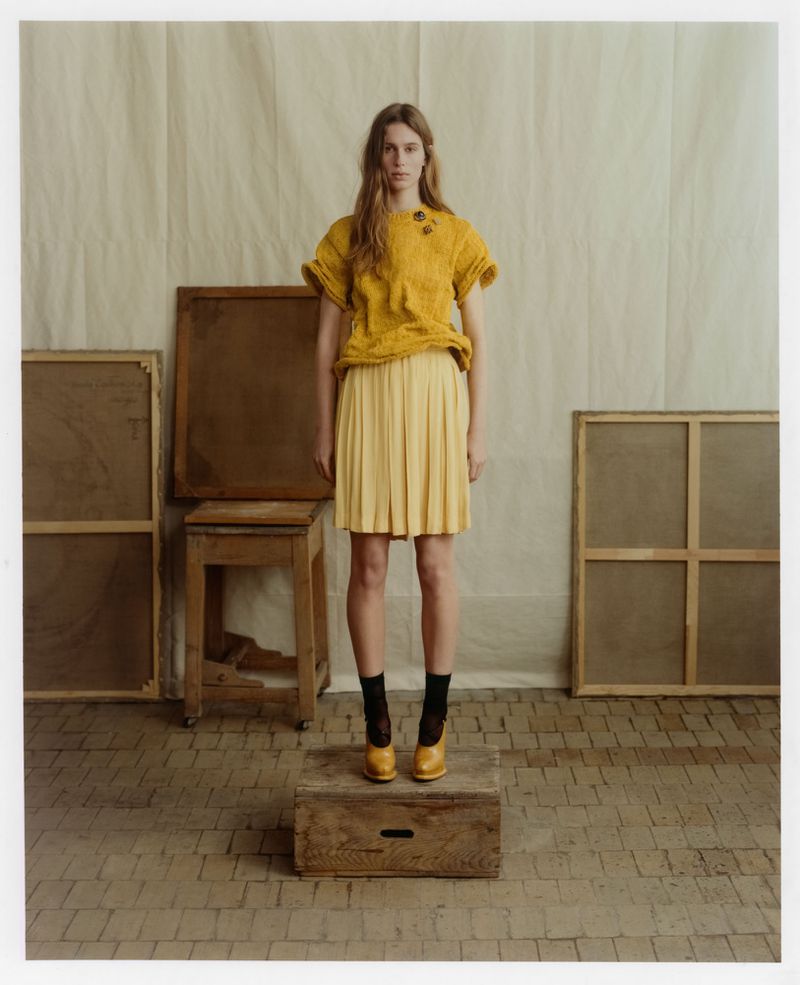 Model standing on a wooden crate in a yellow textured top and pleated skirt against a studio backdrop