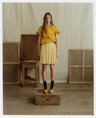 Model standing on a wooden crate in a yellow textured top and pleated skirt against a studio backdrop