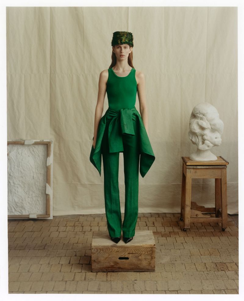 Model standing on a wooden crate wearing a green tank top, green trousers, and a patterned hat against a fabric backdrop.