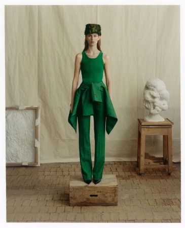 Model standing on a wooden crate wearing a green tank top, green trousers, and a patterned hat against a fabric backdrop.