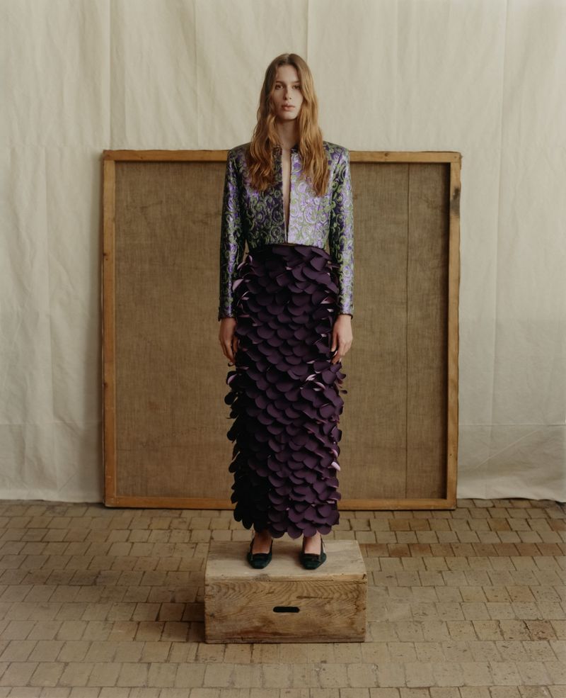 A female model wearing a patterned metallic jacket and textured dark purple skirt standing on a wooden crate