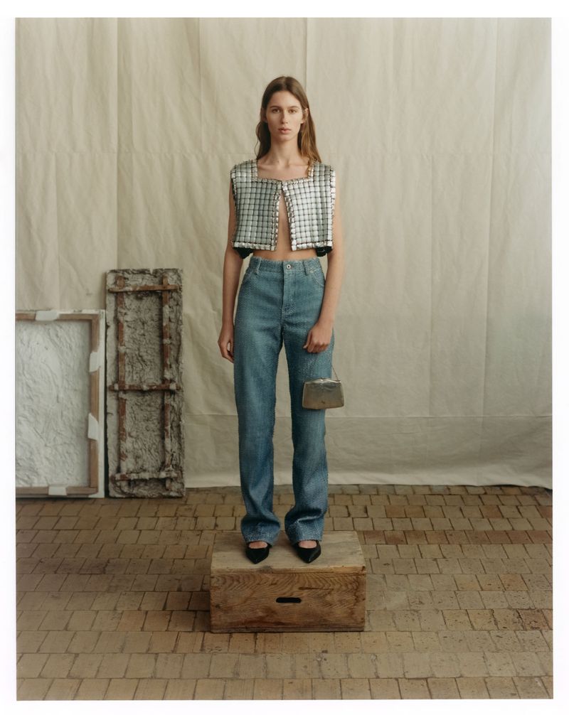 Model standing on a wooden crate wearing a metallic cropped vest and jeans against a textured backdrop