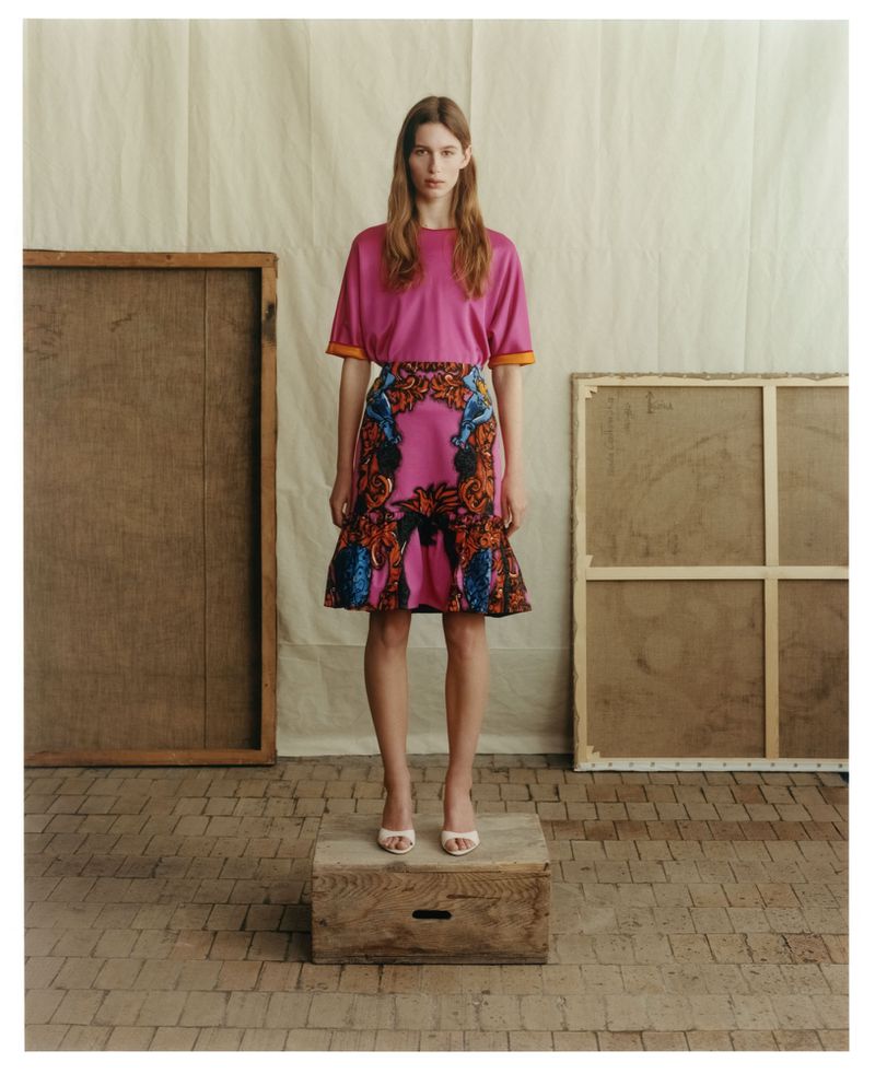 Model standing on a wooden crate wearing a pink top and patterned skirt against a backdrop with canvas frames
