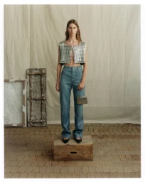 Model standing on a wooden crate wearing a metallic cropped vest and jeans against a textured backdrop