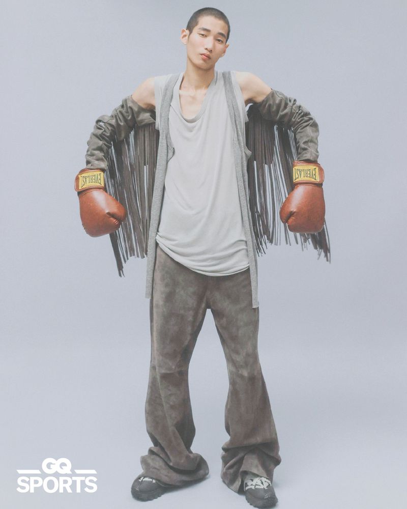 A young male model wearing boxing gloves and fringed sleeves against a neutral grey studio seamless background.