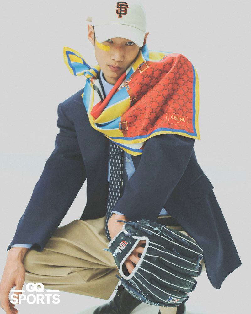 Fashion portrait of a young man wearing a baseball cap, scarf, blazer, and baseball glove against a white backdrop.