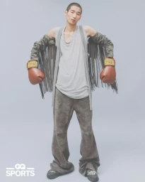A young male model wearing boxing gloves and fringed sleeves against a neutral grey studio seamless background.