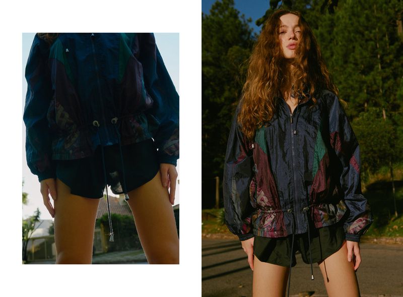 A woman with long wavy hair wearing a patterned windbreaker and dark shorts outdoors against a treed background.