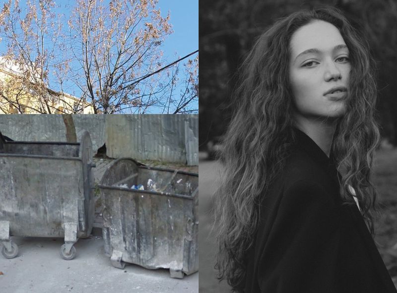 Black and white portrait of a woman with long, curly hair; composition includes a trash bin area and a tree in the background.