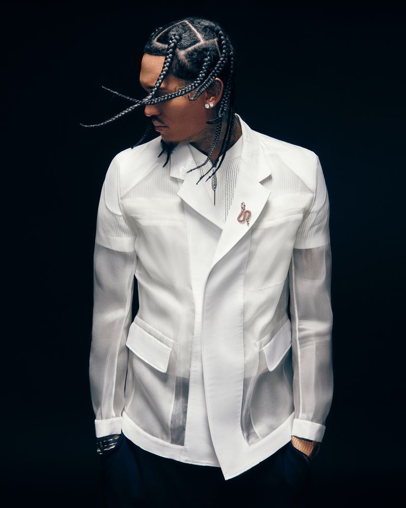 Man with braided hair wearing a white sheer jacket and black trousers against a dark background in a portrait photograph.