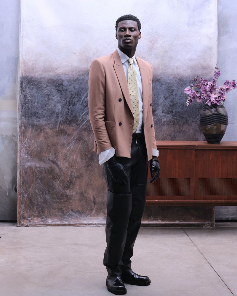 Model wearing a tan blazer, patterned tie, black leather pants, and gloves standing against a textured backdrop for Flanelle Magazine.