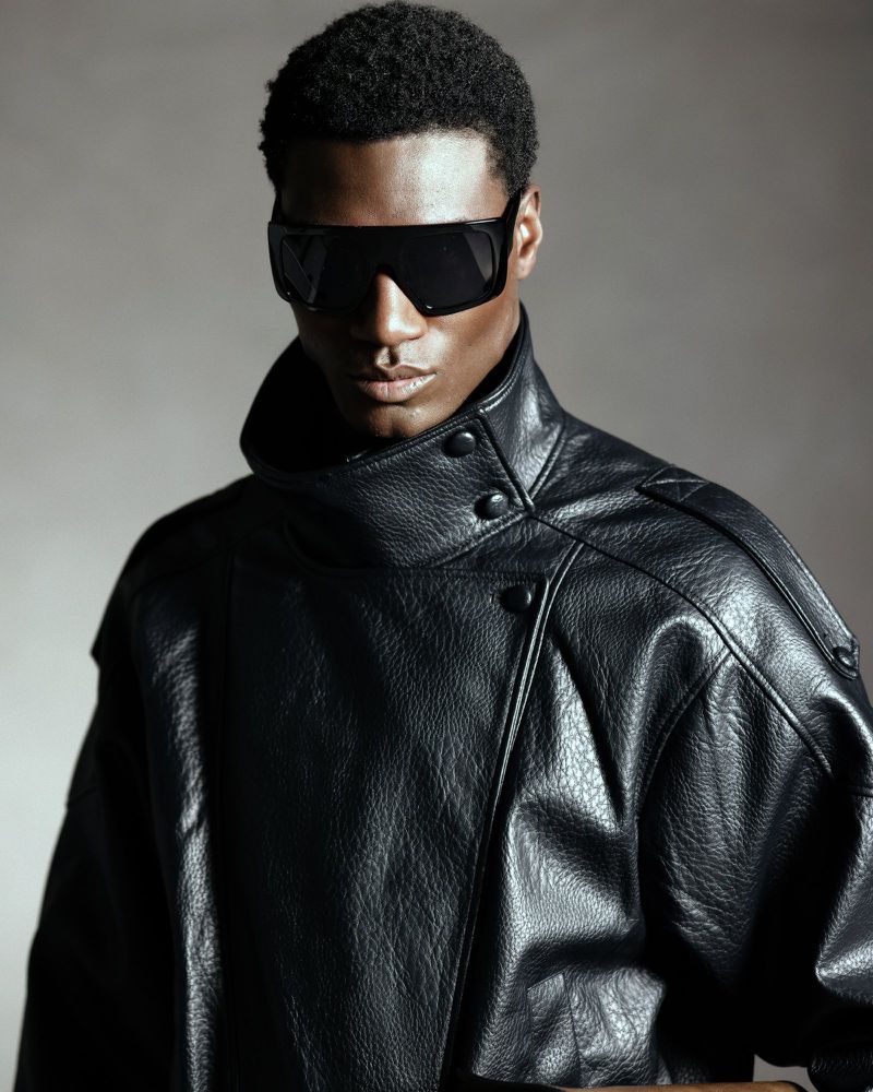 Black man wearing black sunglasses and a high-collared leather jacket against a neutral grey studio backdrop.