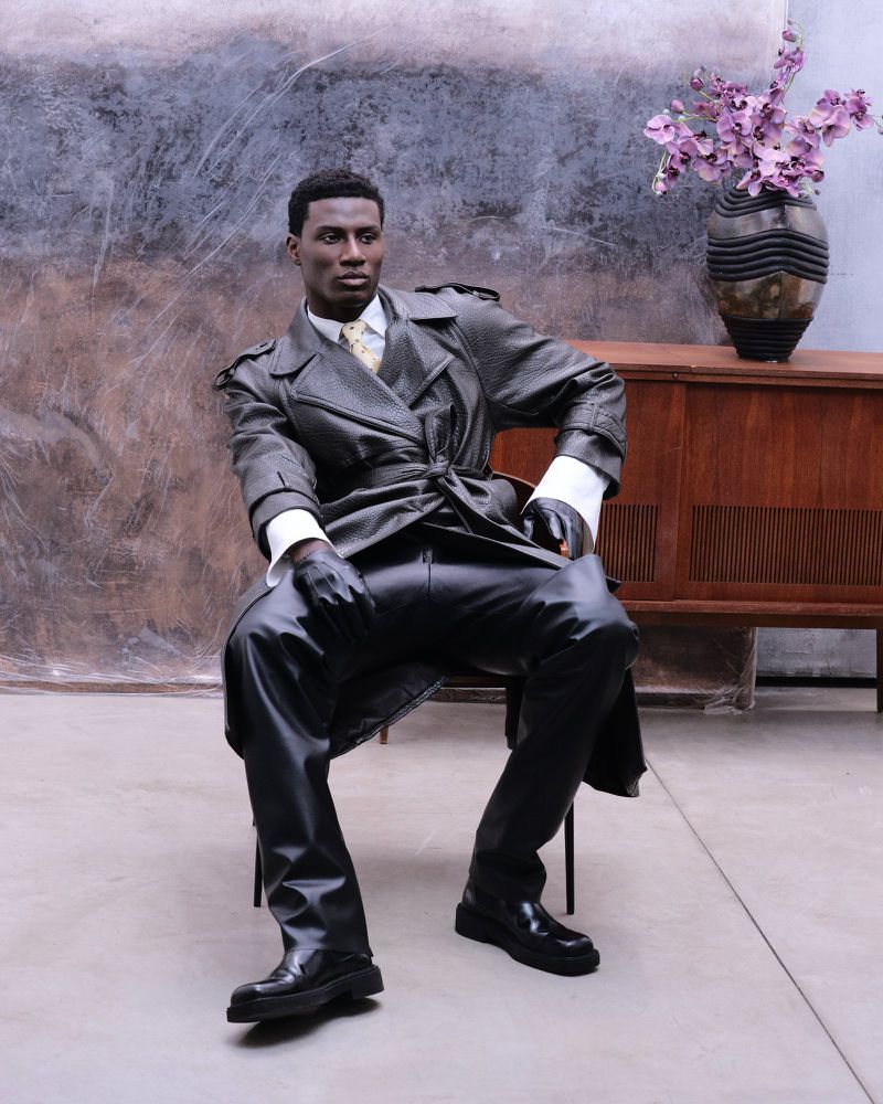 Model seated in a textured trench coat and leather gloves against a concrete wall for Flanelle Magazine