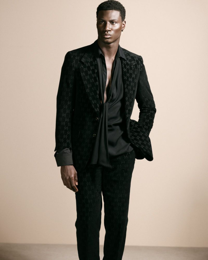 Black man wearing a dark patterned suit and black shirt posing against a neutral warm background for Flanelle Magazine.