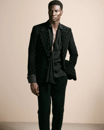 Black man wearing a dark patterned suit and black shirt posing against a neutral warm background for Flanelle Magazine.