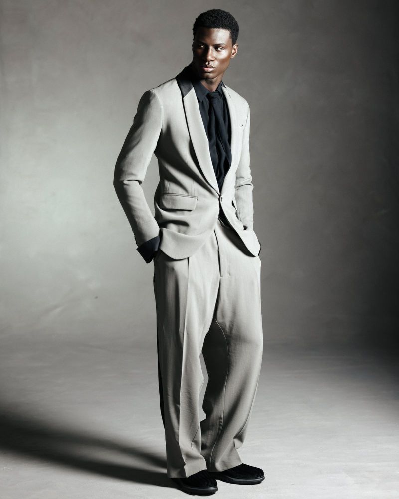 Model wearing a light grey suit with a black shirt and tie posing against a grey studio backdrop for Flanelle Magazine.