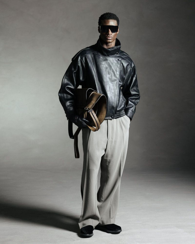 Black man wearing dark sunglasses, black leather jacket and grey trousers holding a bag, against a neutral studio backdrop