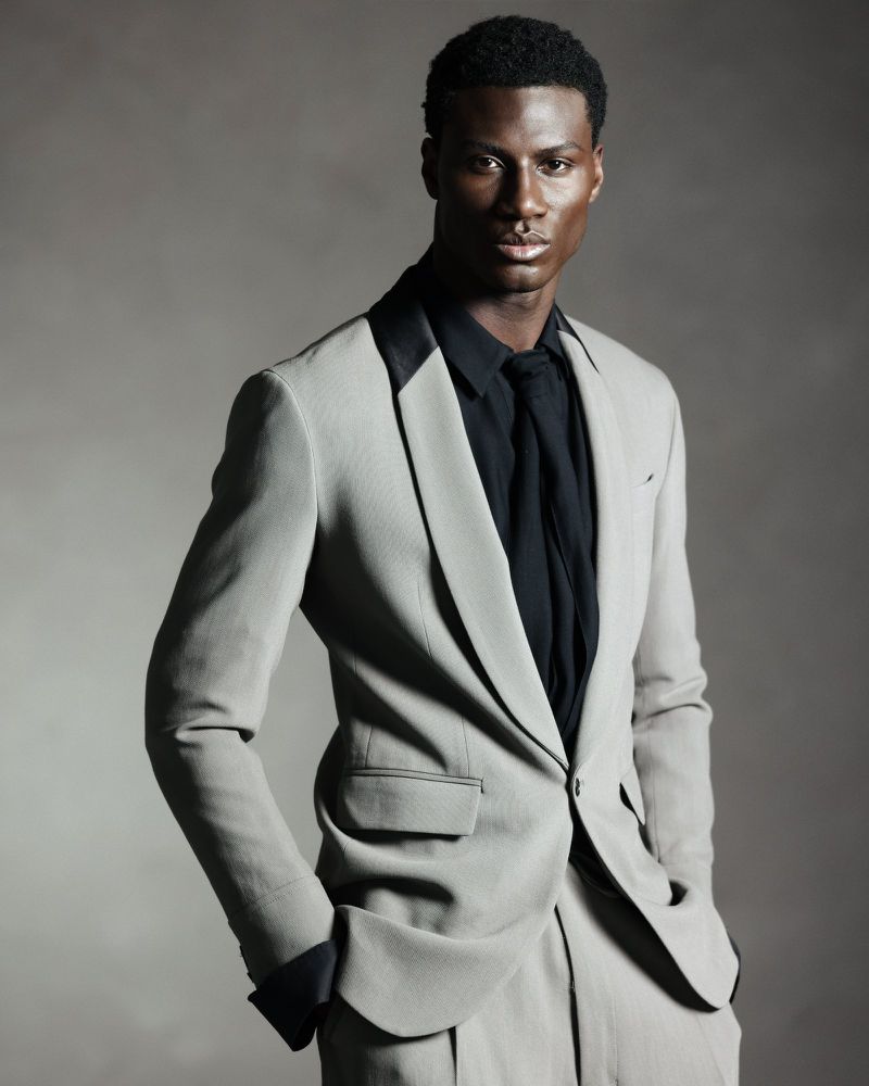 Black man wearing a grey suit jacket and black shirt against a neutral grey studio backdrop