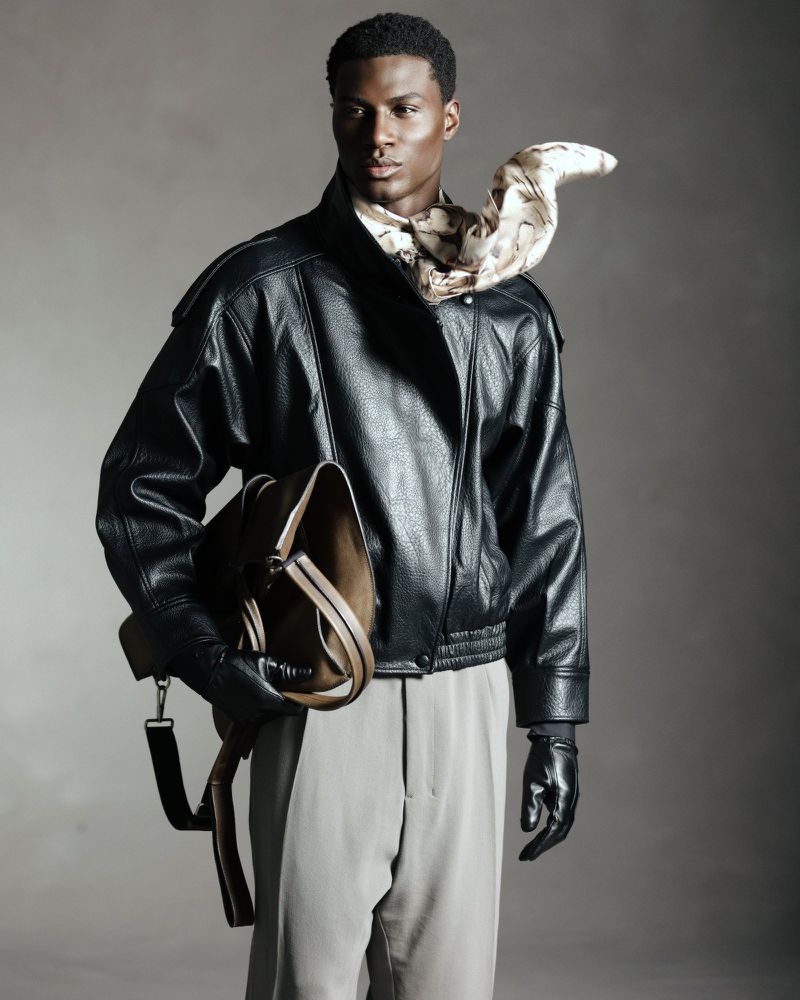 Black man wearing a black leather jacket, grey trousers, and a patterned scarf while holding a brown leather bag
