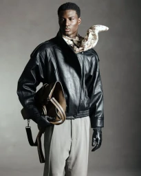 Black man wearing a black leather jacket, grey trousers, and a patterned scarf while holding a brown leather bag