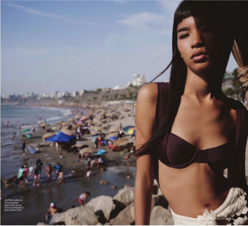 Model in a dark bikini top and white knit skirt posing near a crowded sandy beach under a clear sky.