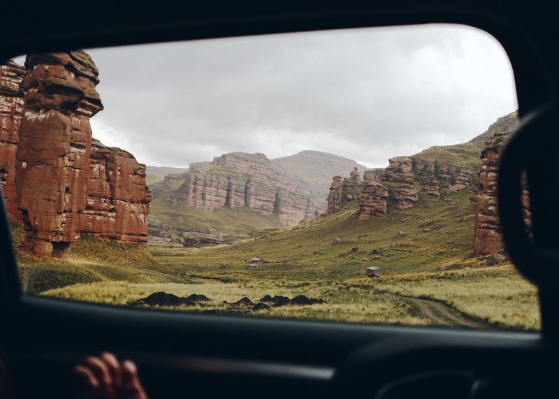 Scenic landscape view through a car window with red rock formations, grassy valley, and cloudy sky, capturing a travel perspective.