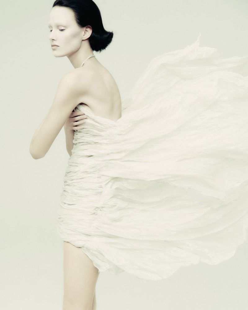 Woman with dark hair and pale skin poses in a textured white dress against a light backdrop in a studio fashion photograph.