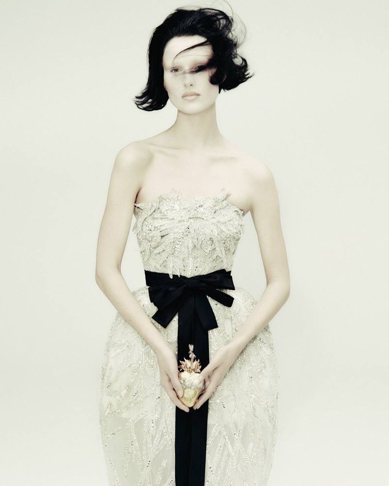 Woman in strapless gown with bow holds decorative object in a studio shot with a light background, showcasing fashion and beauty.